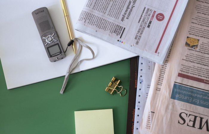 Flat lay of an office desk with newspapers, notepad, and recorder on a green surface.