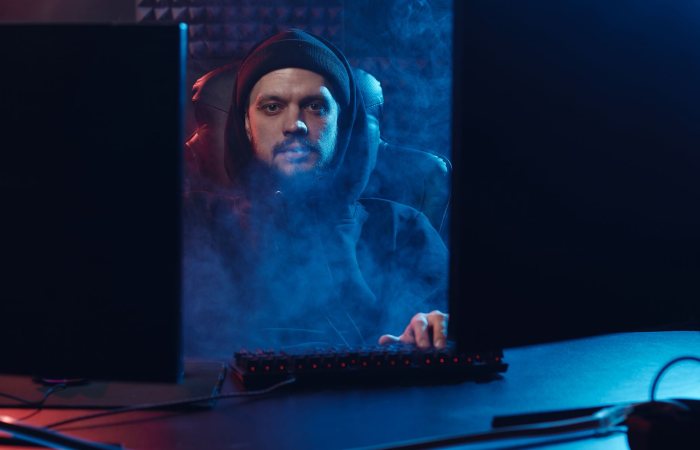 Dramatic scene of an adult man in a hoodie using a computer, surrounded by smoke in a dimly lit room.