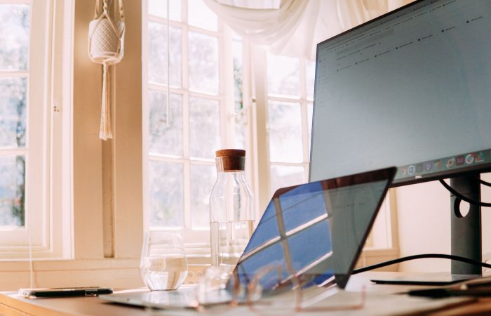 A modern home office setup with a laptop and monitor, bathed in natural light through large windows.