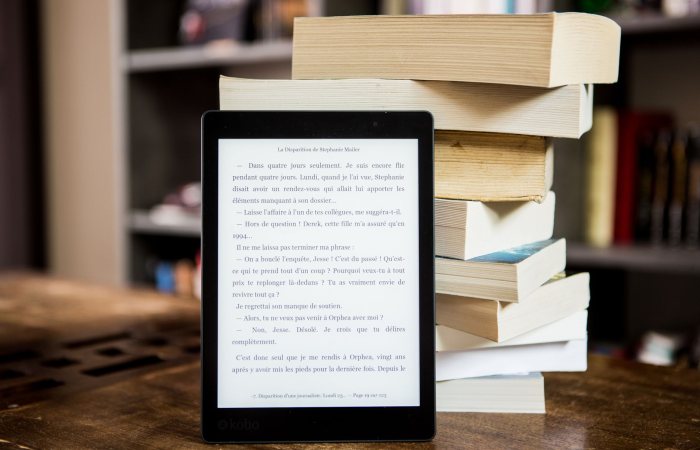 E-reader with text displayed next to a stack of physical books on a wooden table.