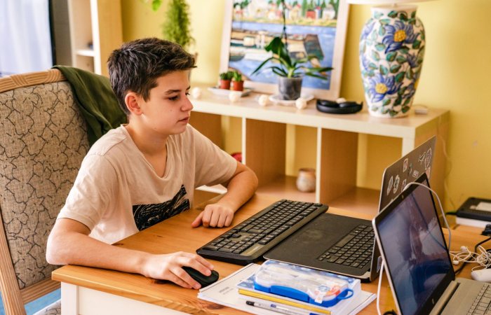 Teenage boy focused on computer work at a home desk setup with dual laptops and keyboard.