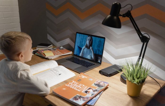 A young boy participates in a virtual class from home, using a laptop and study materials.